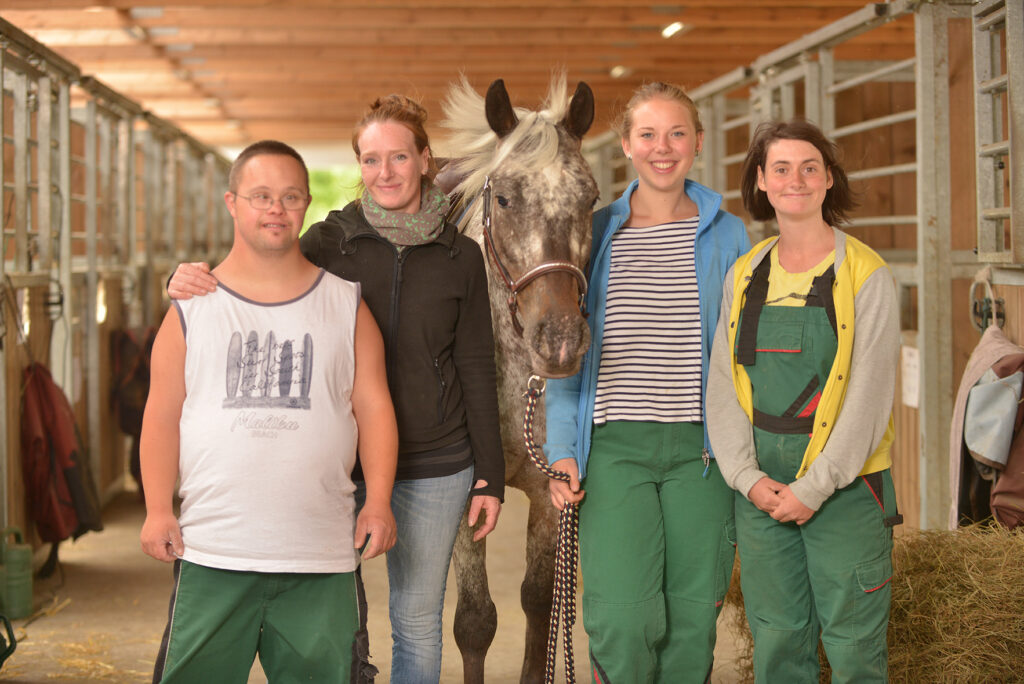 Eine Gruppe von jungen Menschen steht zusammen mit einem Pferd im Stall und lächelt in die Kamera.